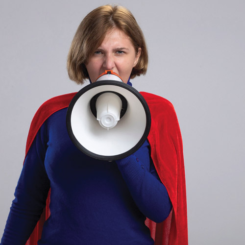 Une femme habillée en bleu qui a une cape rouge sur les épaules. Elle crie dans un mégaphone.
